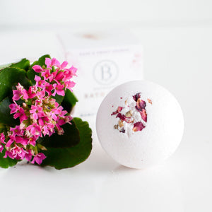 Round white Mama's Perch Bath Bomb | Bathorium with pink flower petals on top, placed beside vibrant pink flowers with green leaves on a white surface.