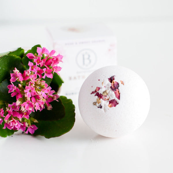 Round white Mama's Perch Bath Bomb | Bathorium with pink flower petals on top, placed beside vibrant pink flowers with green leaves on a white surface.