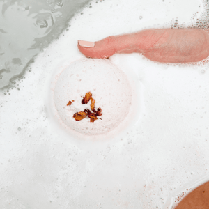 Hand gently holding a foamy, white bath bomb with dried flower petals on its surface, partially submerged in a bath, showcasing Mama's Perch Bath Bomb | Bathorium for a soothing skincare and wellness experience.