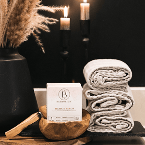 Mama's Perch Bath Bomb | Bathorium displayed in a wooden bowl next to neatly folded beige towels, lit candles, and a black vase with pampas grass, creating a cozy spa setting.