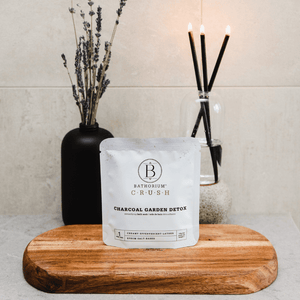 Charcoal Garden Detox Crush | Bathorium bath soak packet displayed on a wooden board with dried lavender in a black vase and lit black reed diffuser candles in a clear glass container in the background, creating a calming spa atmosphere.