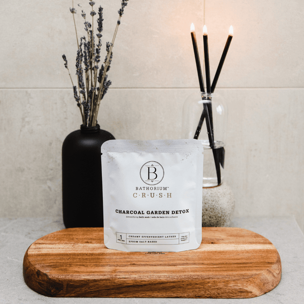 Charcoal Garden Detox Crush | Bathorium bath soak packet displayed on a wooden board with dried lavender in a black vase and lit black reed diffuser candles in a clear glass container in the background, creating a calming spa atmosphere.