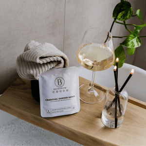 Charcoal Garden Detox Crush | Bathorium bath soak packet on a wooden tray next to a rolled beige towel, a glass of white wine, and a small vase with three lit black incense sticks by a green plant and bathtub.
