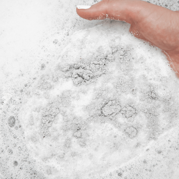 Hand holding and dissolving granular charcoal bath soak in foamy water, showcasing Charcoal Garden Detox Crush | Bathorium for body care and wellness.