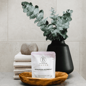 Eucalyptus Apothecary Crush | Bathorium bath soak package displayed on a wooden dish with stacked towels and a black vase holding eucalyptus branches in a spa-like setting.