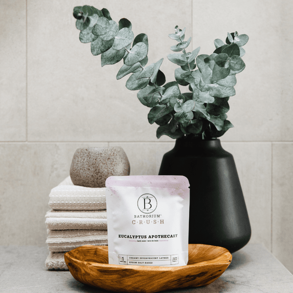 Eucalyptus Apothecary Crush | Bathorium bath soak package displayed on a wooden dish with stacked towels and a black vase holding eucalyptus branches in a spa-like setting.