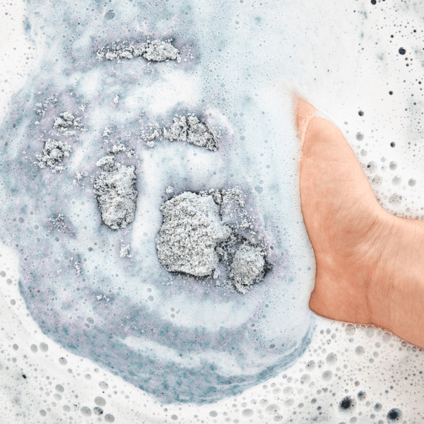 A close-up of a hand swirling a foamy bath soak with shimmering purple and gray tones, showcasing the relaxing texture of Midnight Superbloom Crush | Bathorium.