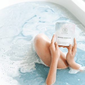 Person relaxing in a bath with light blue water and foam, holding a packet labeled Midnight Superbloom Crush | Bathorium for a soothing skincare and wellness experience.