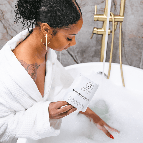 A woman in a white robe holding Northern Sage Recovery Crush | Bathorium bath soak package while relaxing in a bubble-filled bathtub with modern gold fixtures.
