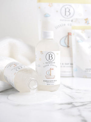 Two bottles of little charlie™ Bubble Bath Milk | Bathorium on a white marble surface, one standing upright and one lying down with liquid spilled beside it, surrounded by soft towels and blurred packaging in the background.