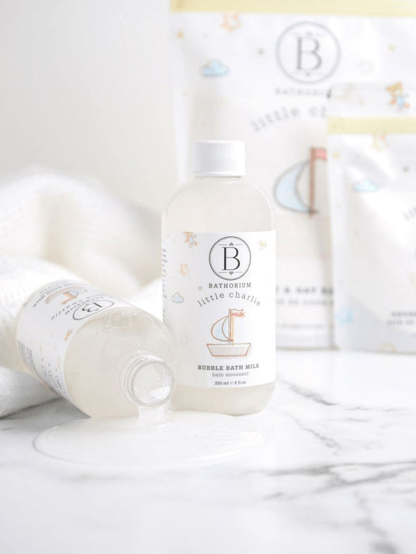 Two bottles of little charlie™ Bubble Bath Milk | Bathorium on a white marble surface, one standing upright and one lying down with liquid spilled beside it, surrounded by soft towels and blurred packaging in the background.