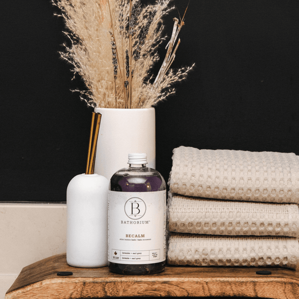 BeCalm Bubble Elixir | Bathorium displayed with a white diffuser, dried pampas grass in a vase, and neatly folded beige textured towels on a wooden surface, promoting calm and wellness.