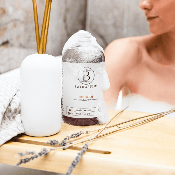 BeCalm Bubble Elixir | Bathorium bottle with foam, placed on a wooden surface next to a white diffuser and lavender sprigs, with a relaxed person in the background enjoying a spa-like setting.