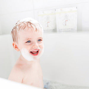 Smiling toddler enjoying a bubbly bath with white foam on head and chin, with two packets of little charlie™ Kids Bath Soak 250g | Bathorium in the background.
