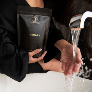 Person holding a black package labeled Hinoki while filling a bathtub faucet with water, promoting relaxation with Hinoki Cypress Magnesium Bath Flakes | Bathorium.