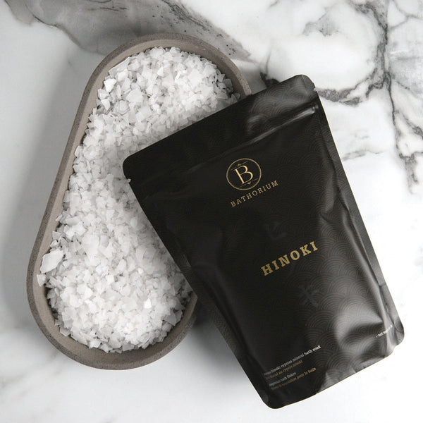 Black resealable pouch of Hinoki Cypress Magnesium Bath Flakes | Bathorium displayed on a marble surface next to a tray filled with coarse white bath flakes for body care and wellness.