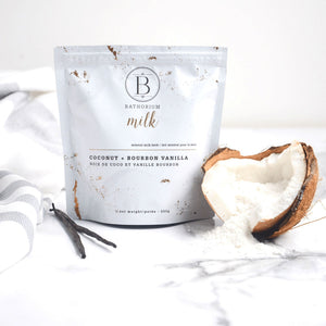 White resealable pouch of Coconut + Vanilla Milk Mineral Soak 650g | Bathorium beside a cracked coconut shell filled with white powder on a marble surface with vanilla pods and a striped towel nearby