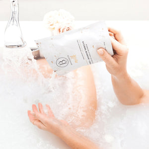 Hand pouring Coconut + Vanilla Milk Mineral Soak 650g | Bathorium into a white bathtub filled with water, with soft flowers in the background for a relaxing spa experience.