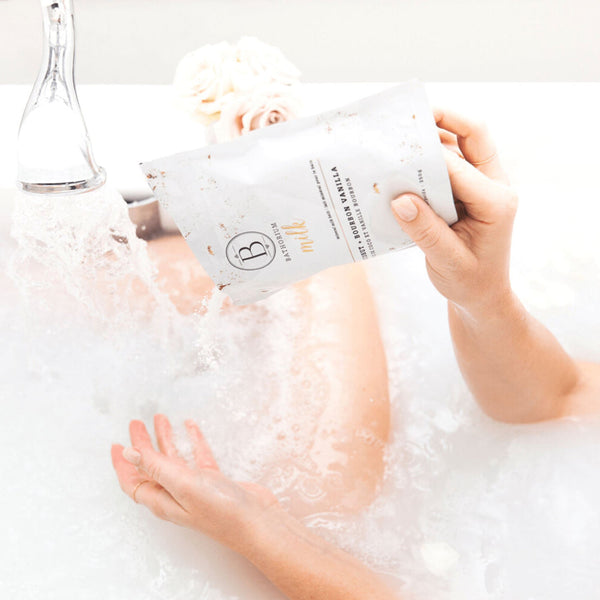 Hand pouring Coconut + Vanilla Milk Mineral Soak 650g | Bathorium into a white bathtub filled with water, with soft flowers in the background for a relaxing spa experience.