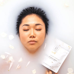 A serene person with eyes closed immersed in a milky bath surrounded by white petals, holding a pack of Coconut + Vanilla Milk Mineral Soak 650g | Bathorium for skincare and wellness relaxation.