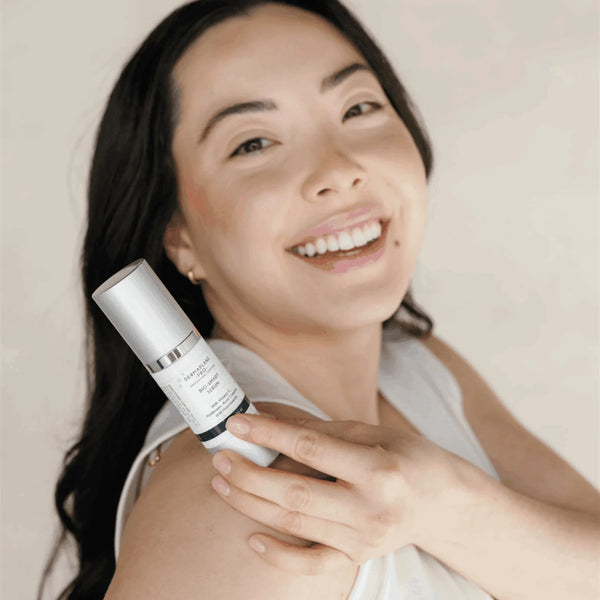 Smiling woman with clear skin holding Bio-Smart Serum | DermaplanePro, showcasing the skincare product in a bright, natural setting promoting wellness.