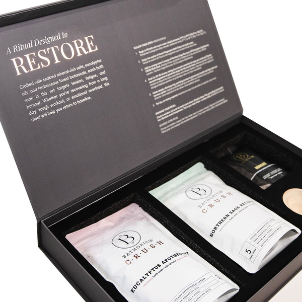 Open gift box set featuring two packets of Bathorium skincare body care products and a small wooden scoop, labeled "Drenched In Renewal Gift Box Set | Bathorium