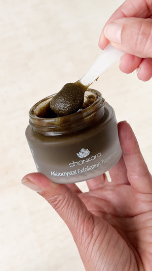 Hand holding an open jar of Microcrystal Exfoliator | Shankara with a small spatula scooping out the dark exfoliating cream, showcasing skincare body care product use.