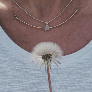Close-up of a person wearing the Diamond Slice Sterling Silver Necklace | Liv & B featuring a delicate silver chain and a small diamond-shaped pendant, paired with a light-colored top and holding a dandelion.