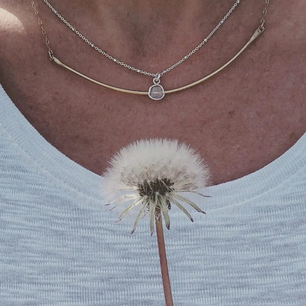 Close-up of a person wearing the Diamond Slice Sterling Silver Necklace | Liv & B featuring a delicate silver chain and a small diamond-shaped pendant, paired with a light-colored top and holding a dandelion.