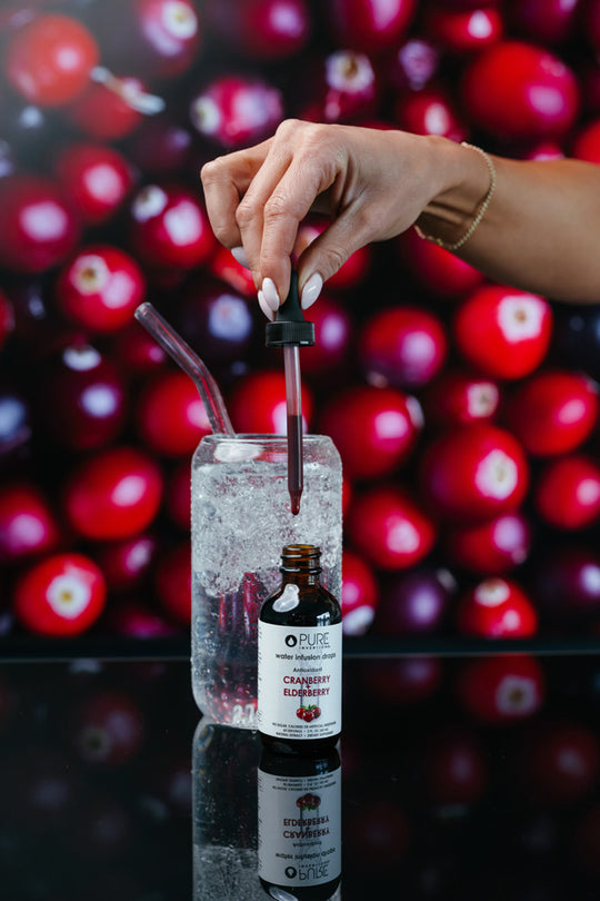 Hand holding dropper with red liquid above a glass of ice water with a straw, next to a bottle of Super Fruit Extracts Water Infusion Drops - Cranberry + Elderberry | Pure Inventions, with a blurred background of fresh cranberries.