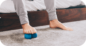 Person using feet to roll a small blue massage tool on carpet for muscle relief and recovery with RAD Roller Soft | RAD.