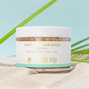 Jar of Coconut Sugar Scrub | Pure Fiji with moringa extract, natural skincare product for body care, placed on beige fabric with green leaves in the background.