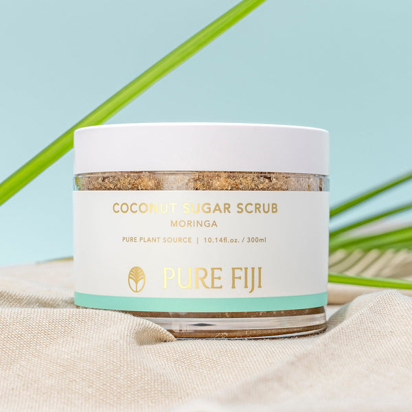 Jar of Coconut Sugar Scrub | Pure Fiji with moringa extract, natural skincare product for body care, placed on beige fabric with green leaves in the background.