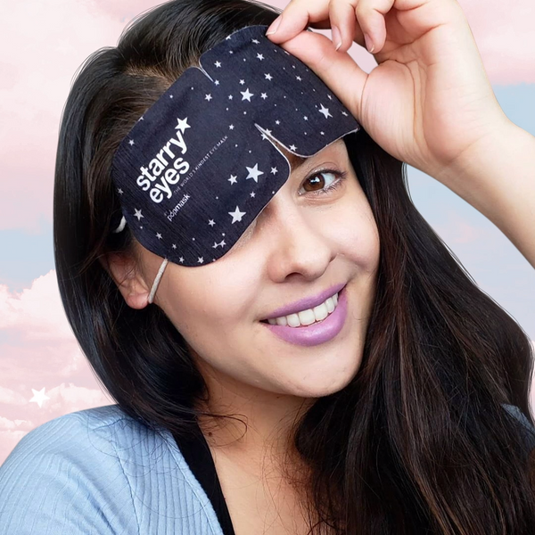 Young woman smiling while wearing a black fabric eye mask with white star patterns labeled Starry Eyes Warming Eye Mask - Single | Popmask against a soft pastel sky background.