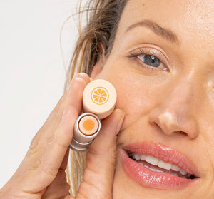 Close-up of a woman holding Orange Mood Fruit™ Lip Therapy | Farmhouse Fresh lip balm near her face, showcasing the product’s orange design and smooth glossy lips.