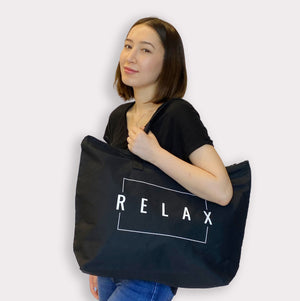 Woman wearing a black t-shirt holding a large black Relax Tote Bag w/ Zipper Top | Lucky Owl with white lettering, styled casually over her shoulder.