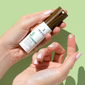 Hand holding a sleek bottle of ORMEDIC balancing eye lift gel | IMAGE Skincare with a wood-grain textured cap against a soft green background, showcasing skincare product for eye care.