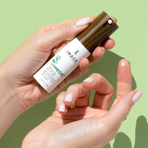 Hand holding a sleek bottle of ORMEDIC balancing eye lift gel | IMAGE Skincare with a wood-grain textured cap against a soft green background, showcasing skincare product for eye care.