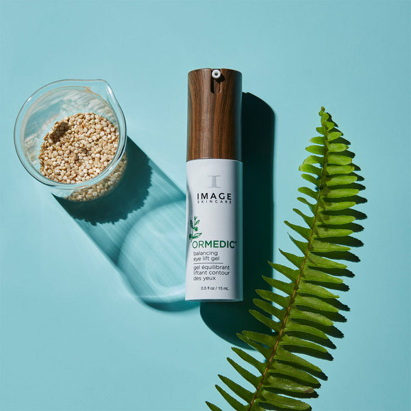 ORMEDIC balancing eye lift gel | IMAGE Skincare bottle with wooden cap next to a green fern leaf and small glass container of natural grains on a blue background, highlighting skincare and wellness.