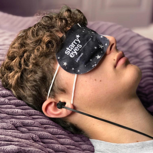 Person lying down with a curly hair wearing the Starry Eyes Warming Eye Mask - Single | Popmask, featuring a dark fabric with star patterns for relaxation and skincare wellness.
