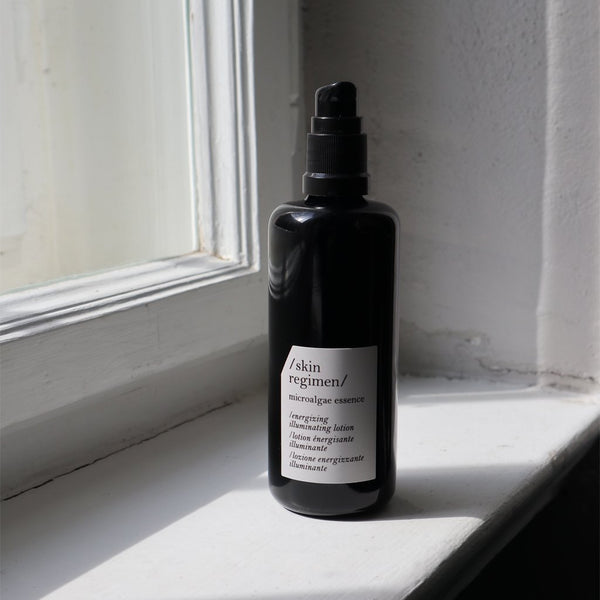 Black pump bottle of Microalgae Essence | /skin regimen/ placed on a white windowsill with natural light highlighting the minimalist skincare product in an elegant setting.