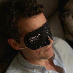 Man relaxing on couch wearing a black Starry Eyes Warming Eye Mask - Single | Popmask with white star patterns for soothing skincare and wellness.